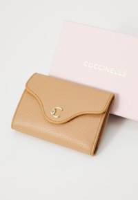 Tan leren portemonnee met een gestructureerde afwerking, gebogen klep en gouden logo-accent. Inclusief een roze doos met "COCCINELLE" in gouden letters.