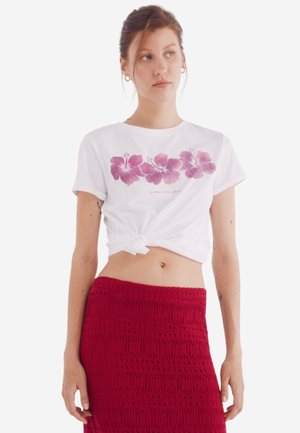 Witte t-shirt met roze hibiscus bloemenprint, geknoopt in de taille; gecombineerd met een rode gehaakte rok met gestructureerde patronen.