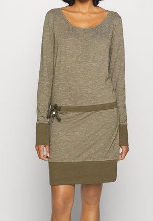 Nainen pukeutuneena oliivinvihreään pitkähihaiseen mekkoon, jossa on vyötäröllä kiristysnauha ja kontrastiväriset kalvosimet sekä helma.