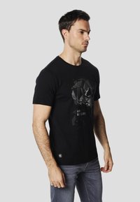 Black cotton T-shirt featuring a geometric design in gray on the front, round neck, and short sleeves. Worn with gray jeans.