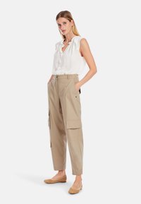 Beige Cargo-Hosen mit Seitentaschen, kombiniert mit einer weißen Bluse mit Rüschchenärmeln. Geflochtene, hellbraune Ballerinas runden das Outfit ab.