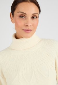 Suéter de cuello alto color crema hecho de una tela suave, con patrones texturizados y un cuello acanalado. Destaca por su apariencia acogedora.