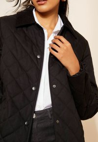 Černá prošívaná bunda s manžetovým límcem, s druky a nařasenými manžetami, nošena přes bílou košili s knoflíky.