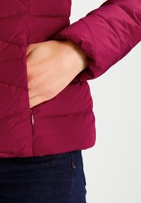 Mano en el bolsillo de una chaqueta acolchada color burdeos, llevada con jeans azul oscuro sobre un fondo claro.