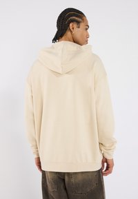 Beige oversized hoodie with a loose fit, featuring a drawstring hood, soft texture, and ribbed cuffs. Paired with dark, worn denim pants.