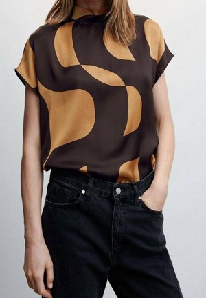 Femme portant un chemisier à manches courtes avec des formes abstraites noires et jaune moutarde rentrée dans un jean noir taille haute, une main dans la poche.