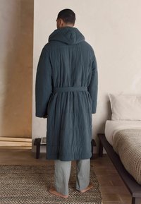 Gestepptes, dunkelblaugrünes Gewand mit Kapuze, aus strukturiertem Stoff und mit Gürtel. Kombiniert mit hellgrauen gestreiften Hosen. Der Boden ist aus Holz.