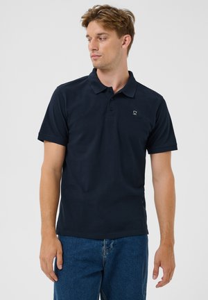 Joven con una camiseta polo negra de manga corta y vaqueros azules, de pie frente a un fondo blanco liso, mirando hacia la izquierda.