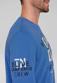 Niebieska koszula z długim rękawem, z nadrukowanym tekstem w białym i czarnym kolorze na przodzie i rękawie, wykonana z teksturowanej bawełny.