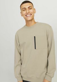 Sweatshirt beige à manches longues, col rond et poche sur la poitrine gauche avec un bord noir. Texture douce, coupe décontractée.