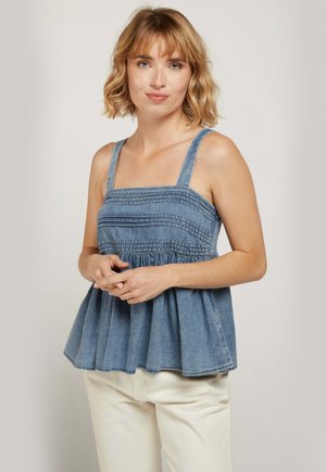 Femme aux cheveux blonds mi-longs portant un haut en denim bleu sans manches et un pantalon blanc cassé, debout les mains jointes de manière détendue.