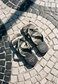 Ζευγάρι σανδάλια Havaianas με καμουφλαρισμένο μοτίβο ακουμπισμένα σε πλακόστρωτο με μαύρα και άσπρα μωσαϊκά με ηλιακό φως και σκιές.