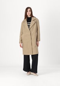 Beige lang frakk med teksturt overflate, krage med utsnitt og sidelommer, båret over en svart og hvit stripete topp og brede svarte bukser.