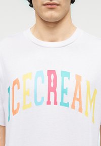 Λευκό βαμβακερό μπλουζάκι με τη λέξη "ICECREAM" σε πολύχρωμα γράμματα. Ο σχεδιασμός χρησιμοποιεί ροζ, μπλε, πορτοκαλί και κίτρινο. Ομαλή υφή.