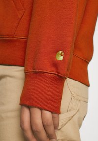 Mano descansando a un lado, vistiendo un sudadera naranja con un pequeño logo amarillo bordado en el puño de la manga, y pantalones beige con un bolsillo visible.