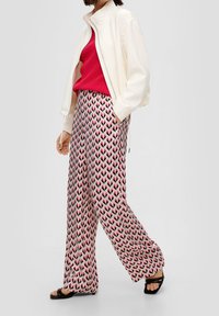 Donna che indossa pantaloni larghi a vita alta con fantasia, top rosso, giacca bianca con zip e sandali neri con punta aperta, in piedi con una mano nella tasca.