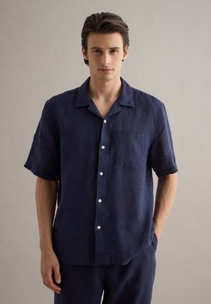 Hombre joven con cabello oscuro, vistiendo una camisa de manga corta azul marino abotonada y pantalones a juego, de pie frente a un fondo beige liso.