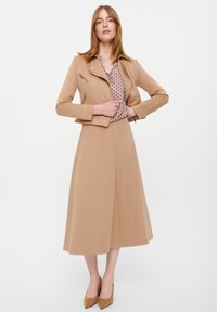 Giacca in pelle cropped color tan sopra una blusa stampata e una gonna midi fluida color tan. L'outfit presenta scarpe con tacco a slip-on e una texture liscia.