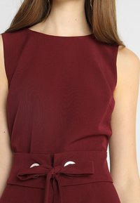 Burgunderfarbenes ärmelloses Top mit rundem Ausschnitt und einer taillierten Taille mit einer gebundenen Schleifendetail. Glattes Material mit matter Oberfläche.