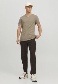 Lys brun t-shirt med korte ærmer og rund halsudskæring, grå bukser og cremefarvede sneakers med maroon accenter. Hvid kasket fuldender outfittet.