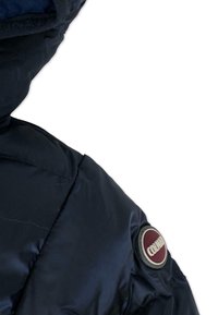 Granatowy pikowany fragment kurtki z zaokrąglonym ramieniem i widoczną naszywką z bordowym okręgiem i białym napisem. Gładka, błyszcząca tekstura.