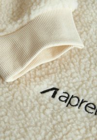 Cremefarvet fleece materiale med en struktureret overflade; har en ribbet manchet og et sort broderet logo på stoffet.