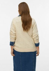 Suéter de punto crema con un diseño de trenzas texturizadas, un cuello redondo y puños acanalados, sobre un vestido de denim con un dobladillo visible.