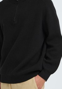 Schwarzer Pullover mit hohem Kragen und halbem Reißverschluss. Hat eine weiche Textur und gerippte Bündchen, kombiniert mit hellbeigen Hosen.