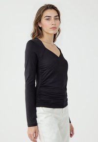 Haut noir à manches longues avec un design enveloppant, présentant des côtés froncés et un décolleté en V profond. Matière lisse avec une silhouette ajustée.