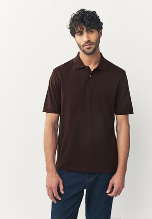 Man met krullend haar en baard, gekleed in een donkerbruin poloshirt en blauwe spijkerbroek, staand tegen een effen lichte achtergrond.