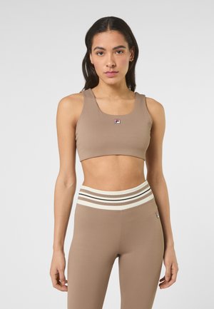 Jeune femme portant un brassard de sport côtelé beige et des leggings taille haute assortis avec une ceinture rayée, se tenant devant un fond uni.
