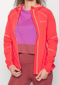 Henkilö, joka pukee kirkasta korallinväristä Columbia-takkia purppuranvärisen toppauksen ja maroonit legginsit päällä, vetää takkia kiinni.