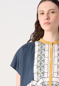 Trumpas rankovės marškiniai su tamsiai mėlynu šonu, raštuota balta dalimi, geltonais mygtukais ir geometriniais gėlių motyvais žaliai geltonoje spalvų schemoje.