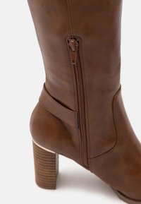 Bota de cuero sintético marrón hasta la rodilla con un cierre lateral, un tacón estructurado y una textura suave. Presenta una correa decorativa cerca del tobillo.