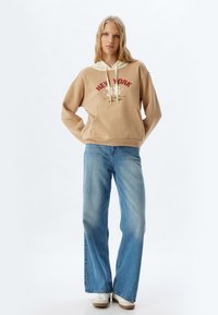 Sudadera beige con capucha forrada en crema y gráfico de "NUEVA YORK", combinada con Jeans de pierna ancha azul claro y zapatillas blancas.