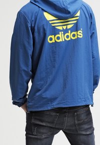 Modrá větrovka s kapucí, s velkým žlutým logem adidas a třemi vodorovnými pásy na zádech. Vyrobena z lehkého materiálu.
