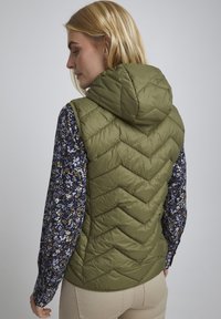 Olivengrøn puffer vest med hætte, prydet med et chevronmønster, lagdelt over en blomstertrykt langærmet skjorte. Glat tekstur, let design.