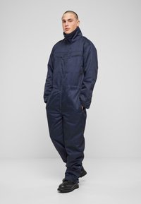 Navy zils kombinezonis, izgatavots no gludas auduma, ar augstu apkakli, divām priekšējām kabatām un elastīgām aprocēm. Valkāts ar melniem izturīgiem zābakiem.