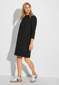 Schwarzes, strukturiertes Kleid mit Dreiviertelärmeln und einer Kordelzugkapuze, getragen mit weißen Sneakers mit schwarzen Akzenten und einer braunen Sohle.