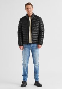 Schwarze Steppjacke mit ge quilteter Textur, hohem Kragen und Reißverschluss. Getragen über einem cremefarbenen Pullover und kombiniert mit hellblauen Jeans und schwarzen Stiefeln.