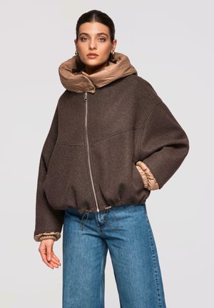 Kobieta w oversize'owej brązowej kurtce zapinanej na zamek z pikowanym kołnierzem i mankietami, w połączeniu z niebieskimi dżinsami, stojąca na jednolitym tle.