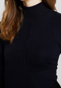 Haut à col roulé côtelé bleu marine avec panneaux texturés et détail de couture centrale. Tissu en maille douce, silhouette ajustée et design à col haut.