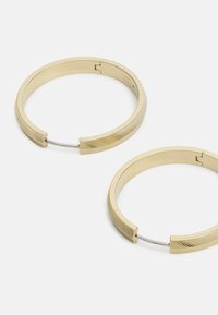 Boucles d'oreilles créoles en métal doré avec une surface lisse et des accents texturés. Le design comprend un fermoir sécurisé et une forme épurée et minimaliste.