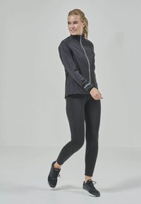 Svart idrottsjacka med hög krage och kontrasterande vit dragkedja, kombinerad med svarta leggings och svarta träningsskor, mot en ljus bakgrund.