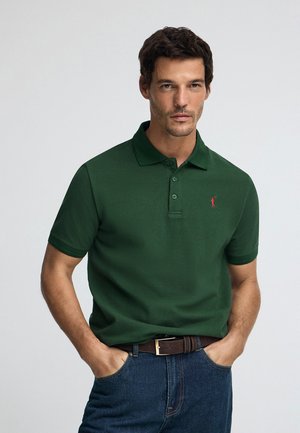 Polo verde in un design classico con colletto e tre bottoni, con un piccolo logo rosso ricamato sul petto a sinistra. Jeans di denim.