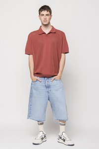 Polo de culoare roșu ruginit, cu mânecă scurtă și un model texturat; asortat cu șorturi din denim de lungime până la genunchi, de culoare albastră deschisă. Adidași albi și negri, cu șosete în dungi.