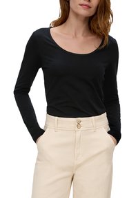 Schwarzes Langarmoberteil mit Rundhalsausschnitt, kombiniert mit cremefarbenen, hochgeschnittenen Hosen mit zwei Knöpfen und seitlichen Taschen.