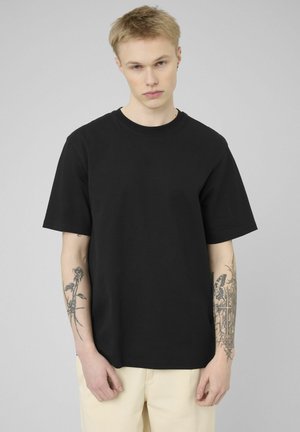 Jeune homme aux cheveux blonds courts portant un t-shirt noir uni et un pantalon clair, montrant des tatouages sur les deux avant-bras sur un fond gris.