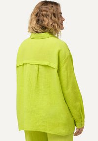 Donna con i capelli biondi mossi, che indossa una maglietta verde lime morbida e strutturata, insieme a pantaloni coordinati, di spalle rispetto a uno sfondo semplice.