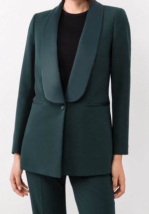 Vrouw draagt een donker groene op maat gemaakte blazer met satijnen sjaalkraag en bijpassende broek, gecombineerd met een zwart geribd topje eronder.